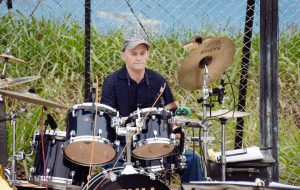 Man playing a drum set outdoors in front of a chain-link fence with greenery in the background.